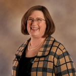 Lori_cropped A person with short, light brown hair wearing a beige and black plaid jacket over a black top, accessorized with a pearl necklace, posed against a softly blurred brown and beige background.